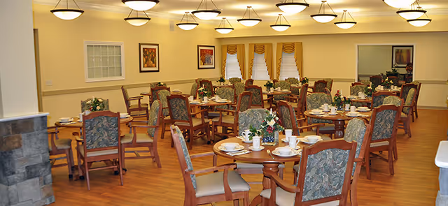 A spacious senior living dining room with round tables and floral centerpieces.