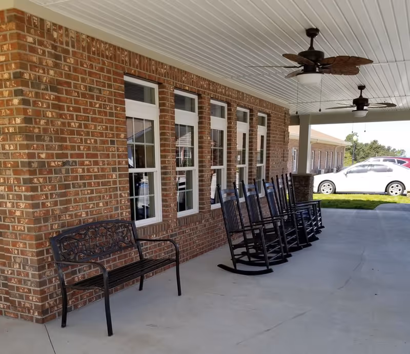 Covered outdoor patio area at Habersham Retreat with a row of black rocking chairs and a black metal bench against a brick wall with multiple windows. Two ceiling fans with leaf-shaped blades hang from the white ceiling. A white car is parked on the driveway nearby.