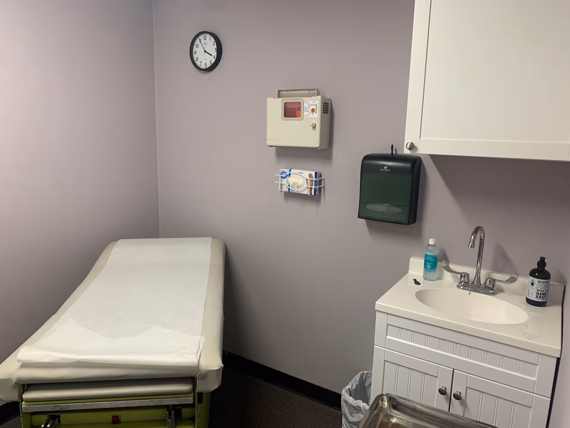 Examination room with a treatment bed, clock, sink, and medical supplies.