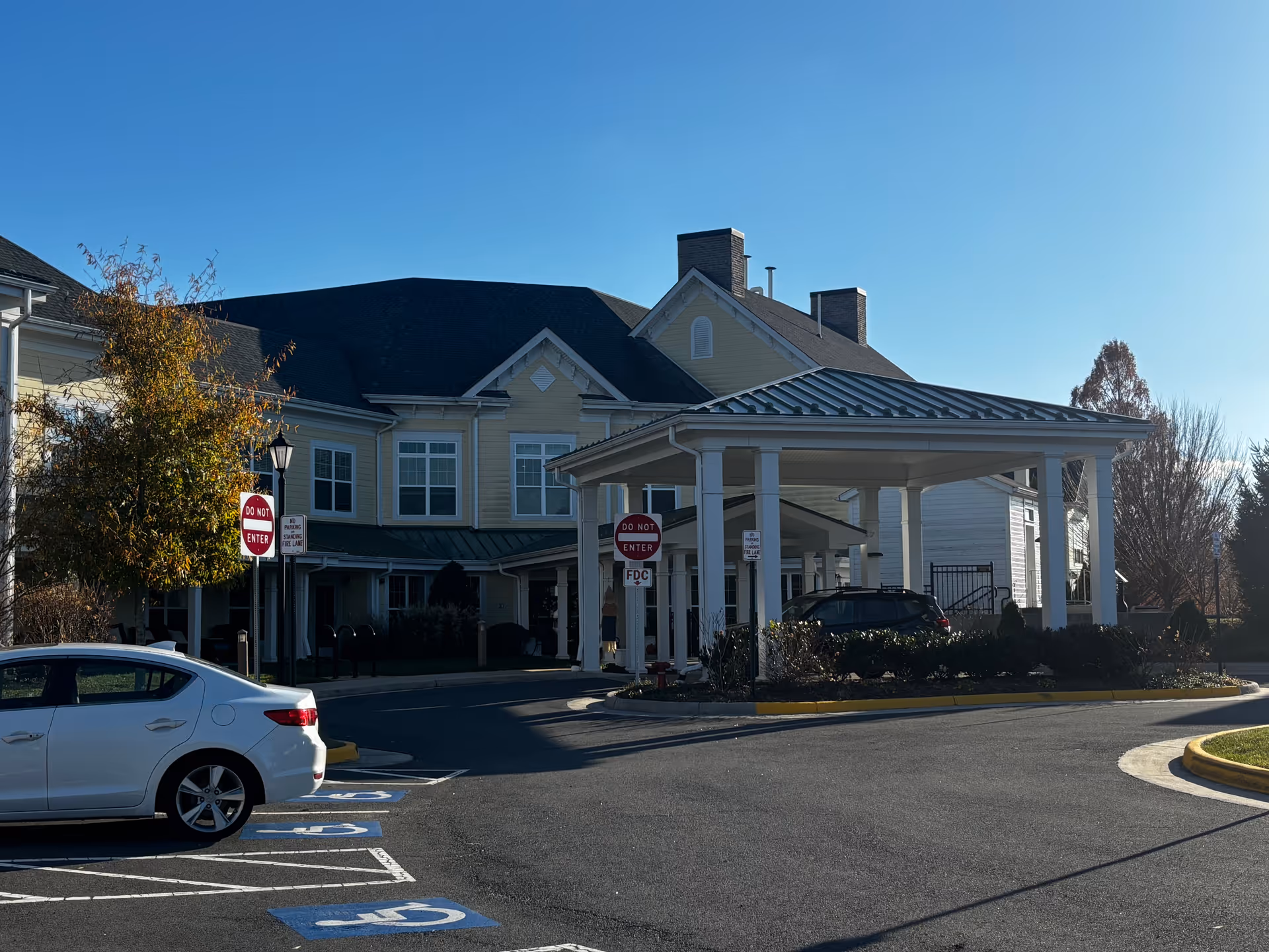 Exterior view of Sunrise at Silas Burke House showing the front entrance with a covered drop-off area, a white car parked in a handicapped parking space, and clear blue sky.