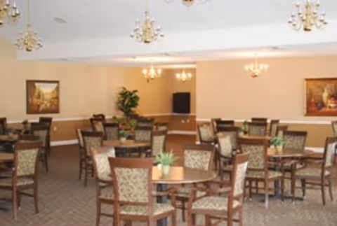A spacious dining room with multiple round wooden tables surrounded by patterned cushioned chairs. The room is decorated with framed paintings on the walls, potted plants, and elegant chandeliers hanging from the ceiling. The walls are painted in a warm beige tone with white trim.