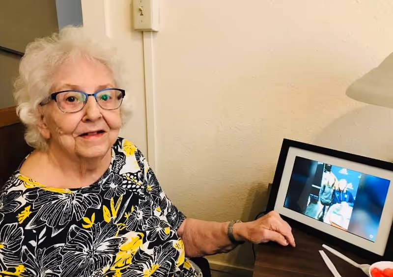 An elderly woman with white hair and glasses is sitting indoors next to a table. She is wearing a black, white, and yellow floral patterned top and smiling slightly. On the table beside her is a digital photo frame displaying a picture of two people. A lamp is partially visible on the right side of the table.