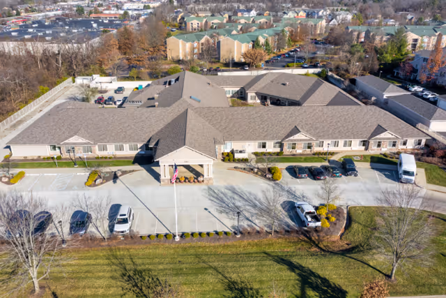 Aerial view of a single-story senior living facility building with a large parking lot in front, surrounded by trees and residential buildings in the background. The building has a beige exterior with a gray roof and a covered entrance with an American flag.