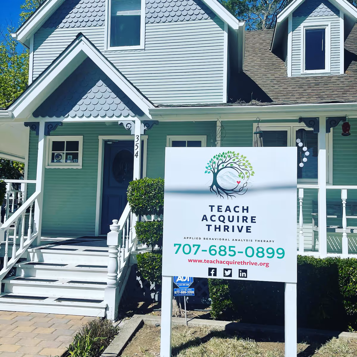 Pastel green house exterior with a large Teach Acquire Thrive sign in the front yard.
