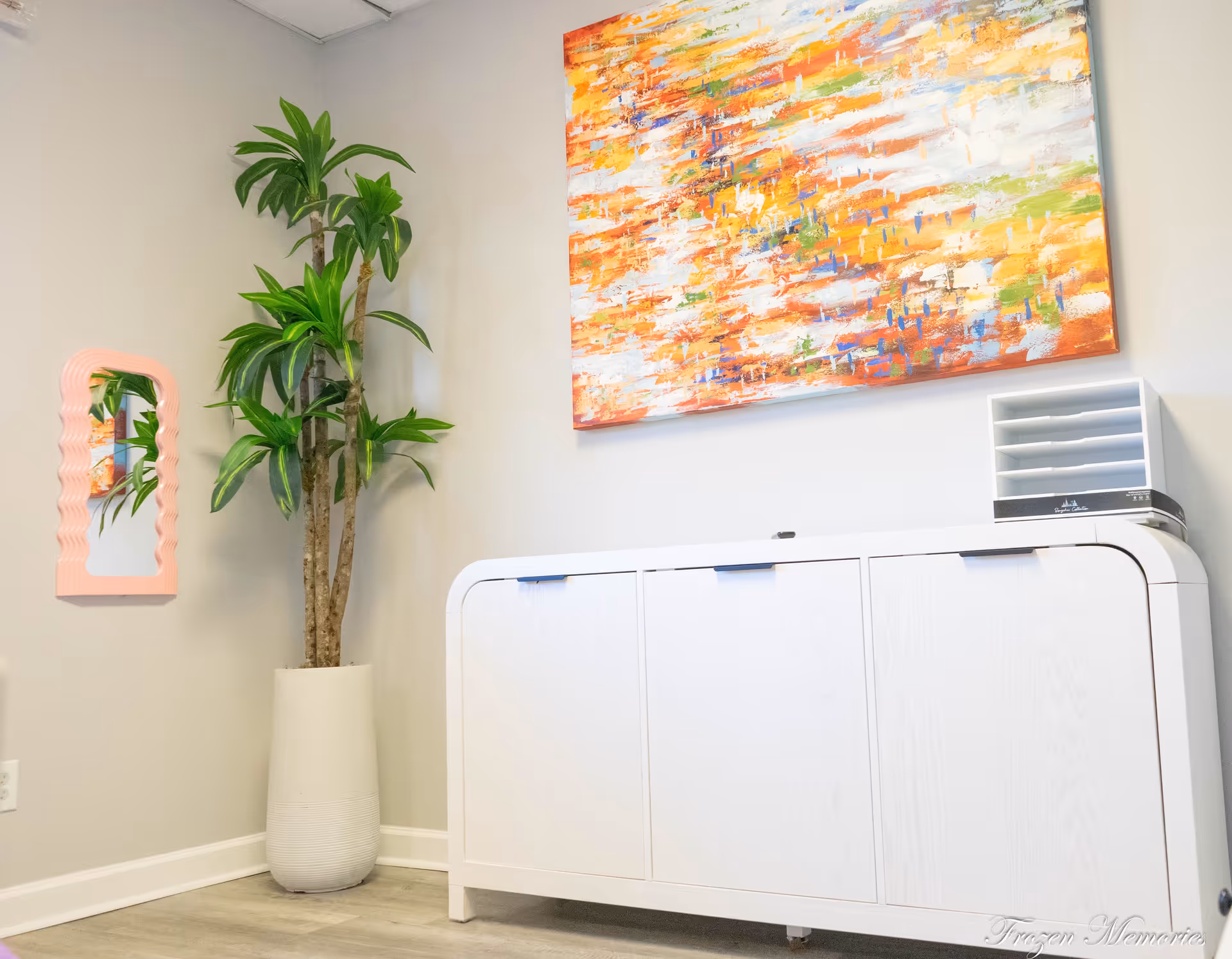 Bright interior area with a large colorful painting, tall plant, and white cabinet.