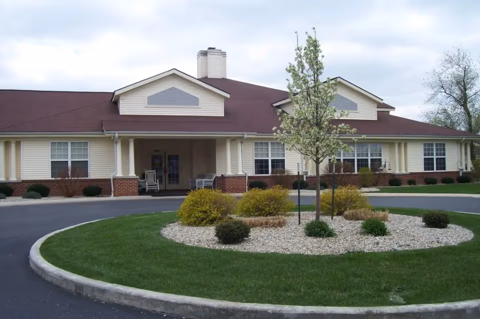 Front exterior view of Bethany Pointe Health Campus showing a single-story building with beige siding and a brown roof. There is a circular driveway with a landscaped island in the center featuring a small tree and shrubs. The entrance has a covered porch with chairs.