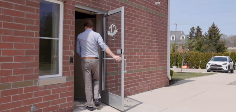 Man entering a brick hospice house through a glass door; sidewalk and car visible outside.