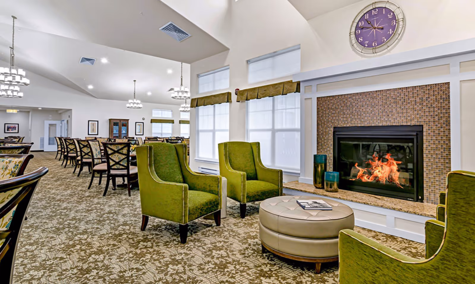 Spacious communal dining and lounge area with green armchairs, a round ottoman, a fireplace, and multiple dining tables.
