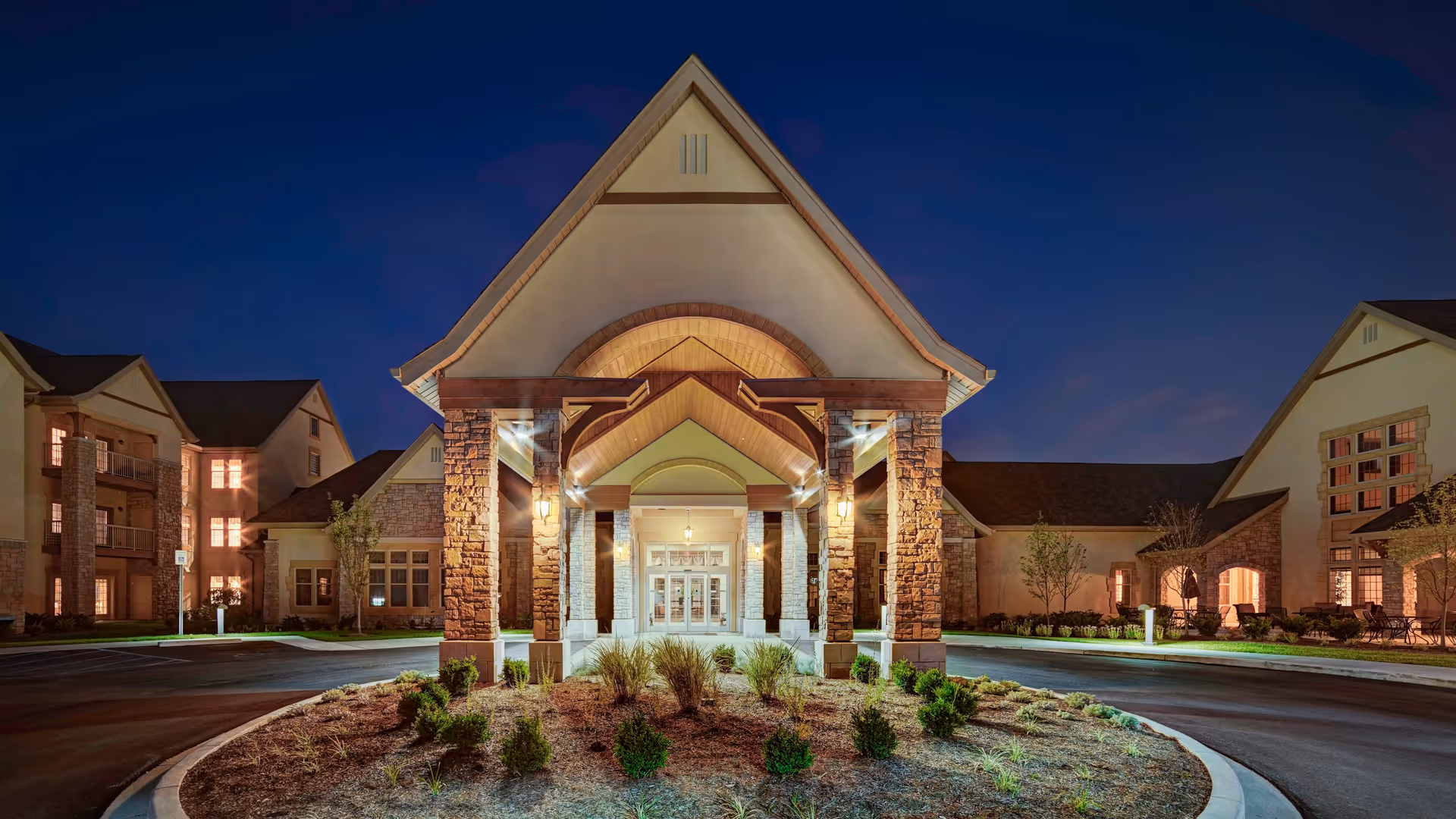 Night view of the front entrance of a senior living facility with a large covered driveway supported by stone pillars, surrounded by landscaped bushes and plants, and illuminated by warm exterior lights.