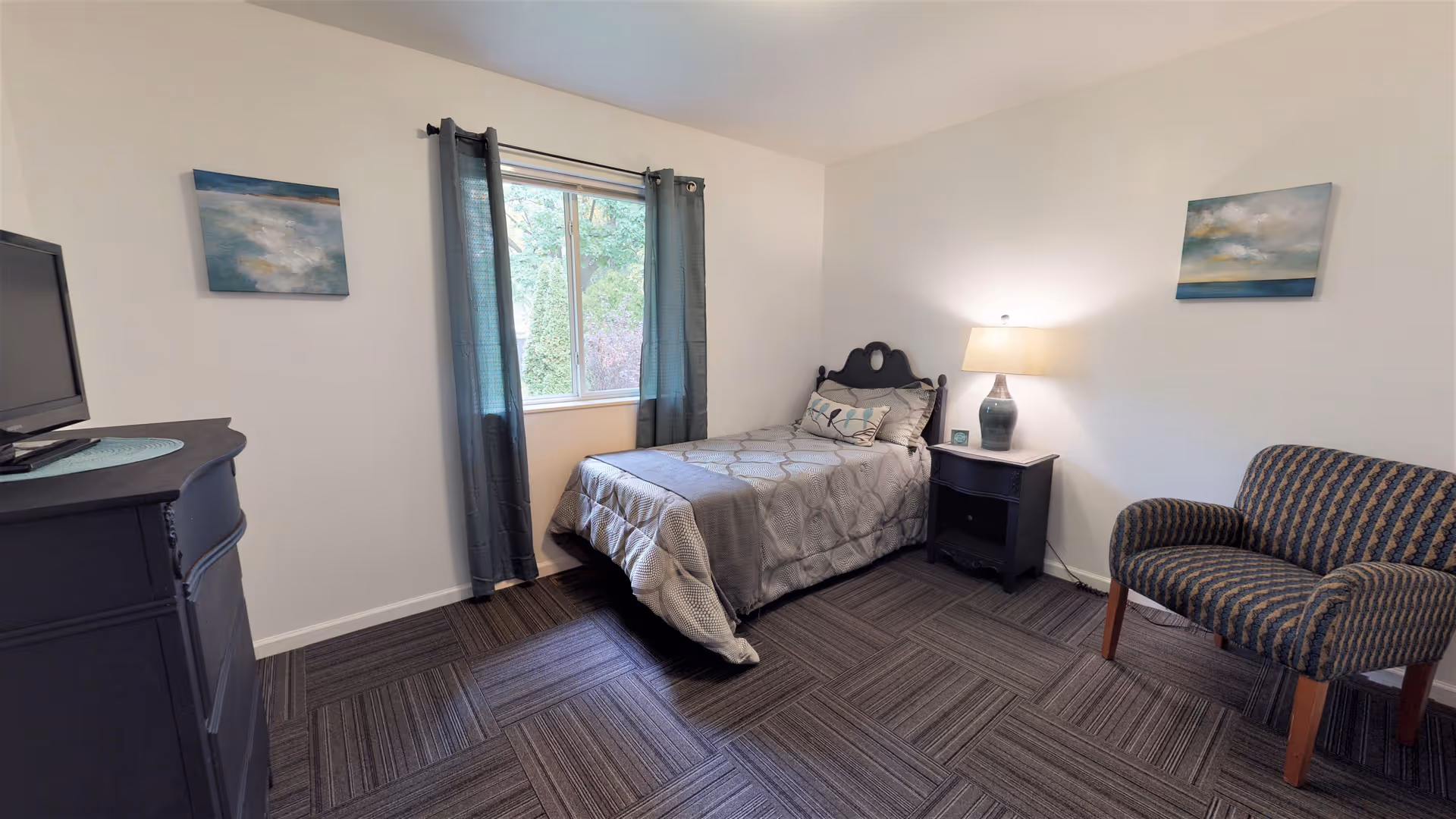 A small, neatly arranged bedroom with a single bed covered in a gray patterned comforter, a nightstand with a lamp, a cushioned armchair with a striped pattern, a dresser with a TV on top, and a window with dark curtains letting in natural light. Two abstract paintings hang on the white walls.