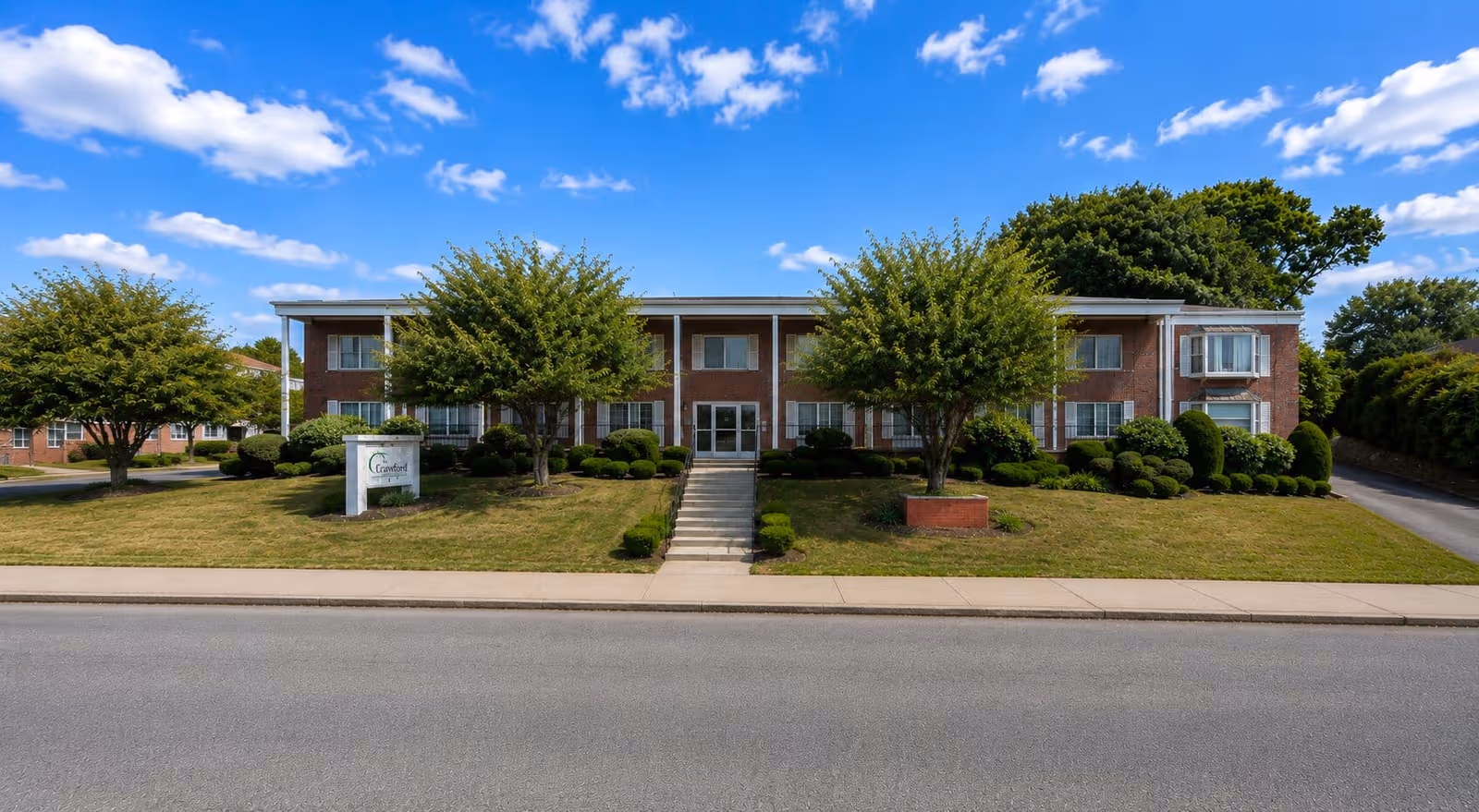 Brick two-story senior living center with manicured lawn, trees, and a sidewalk entrance.