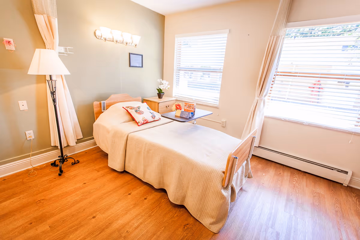 A bright and clean single bedroom in a skilled nursing facility with a wooden bed covered in a beige blanket and a butterfly-patterned pillow. Next to the bed is a small wooden nightstand with a potted plant and a small table with flowers and informational cards. The room has wooden flooring, a floor lamp, and two large windows with white blinds and curtains letting in natural light.