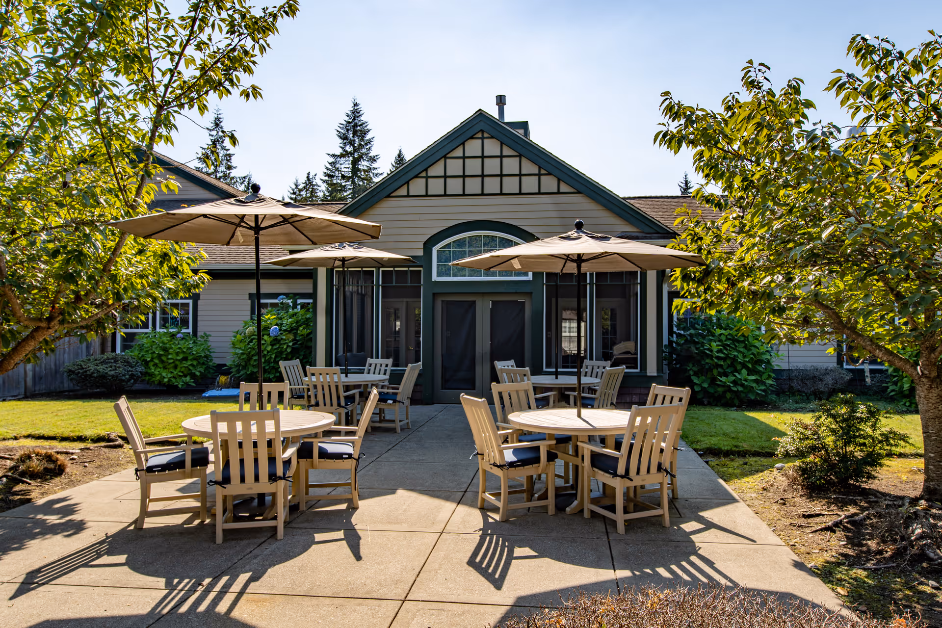 Outdoor patio area at The Ridge an Encore Community featuring round tables with umbrellas and wooden chairs, surrounded by trees and greenery, with a building entrance in the background.