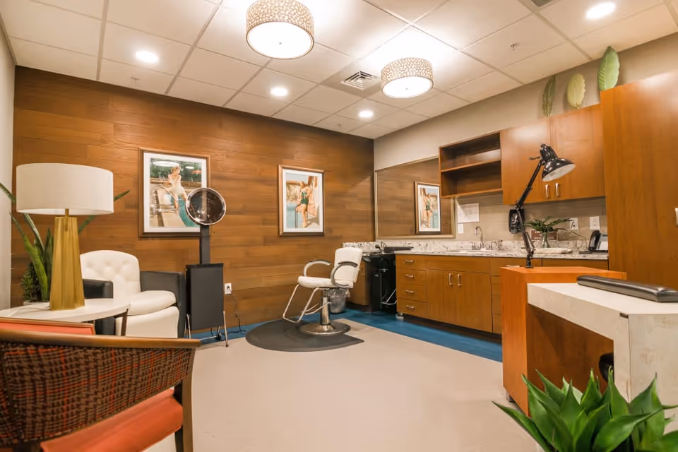 A well-lit interior room with a salon chair in front of a wooden wall adorned with two framed pictures. The room features a white cushioned chair, a floor lamp with a white shade, a small table, and a cabinet with a sink and countertop. There is also a desk lamp, some plants, and a large mirror on the wall.