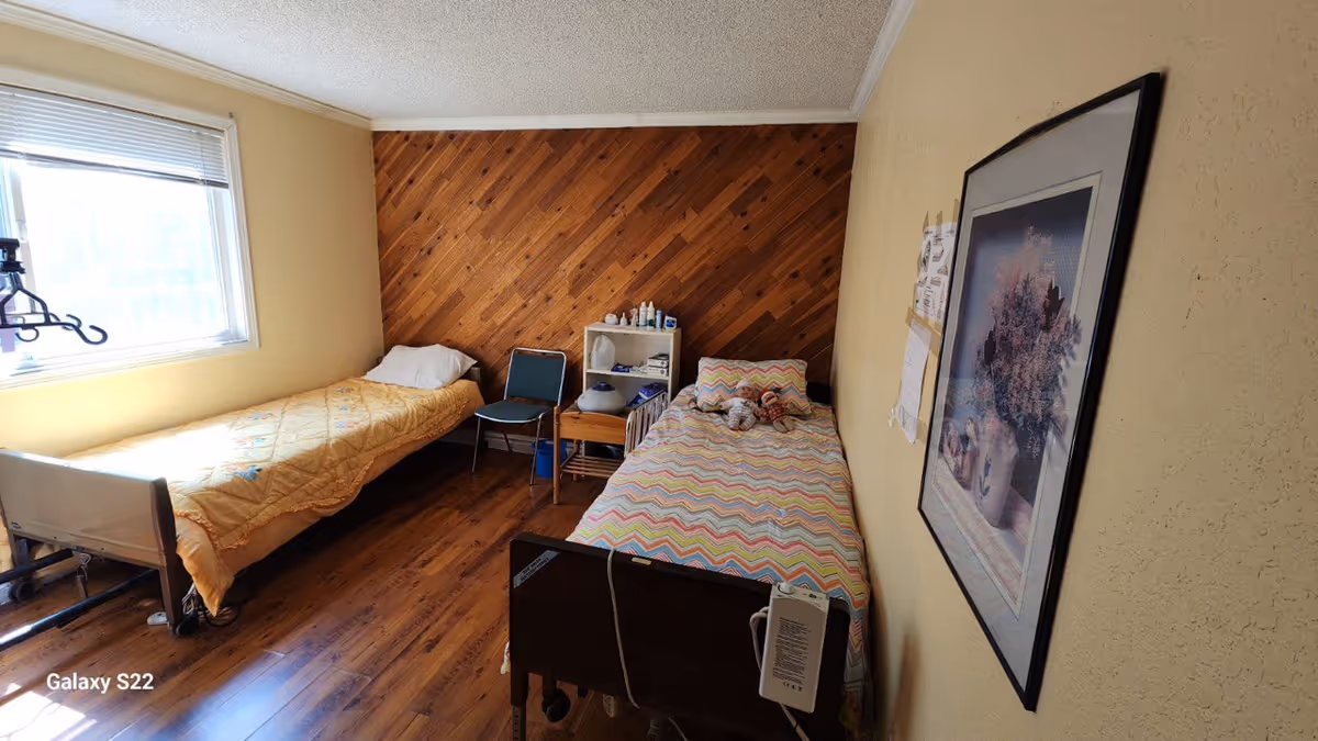 Two single beds in a small beige bedroom with wood panel accent wall, window, and chair.