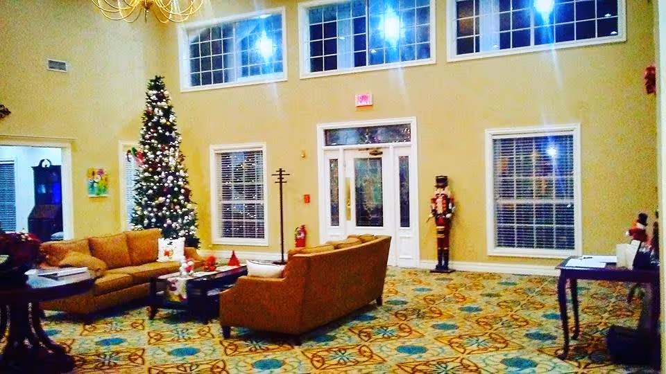 A spacious living room area decorated for Christmas with a tall Christmas tree adorned with lights and ornaments. The room features two brown sofas arranged around a coffee table, a patterned carpet, large windows with blinds, a chandelier, and a nutcracker statue near the exit door.