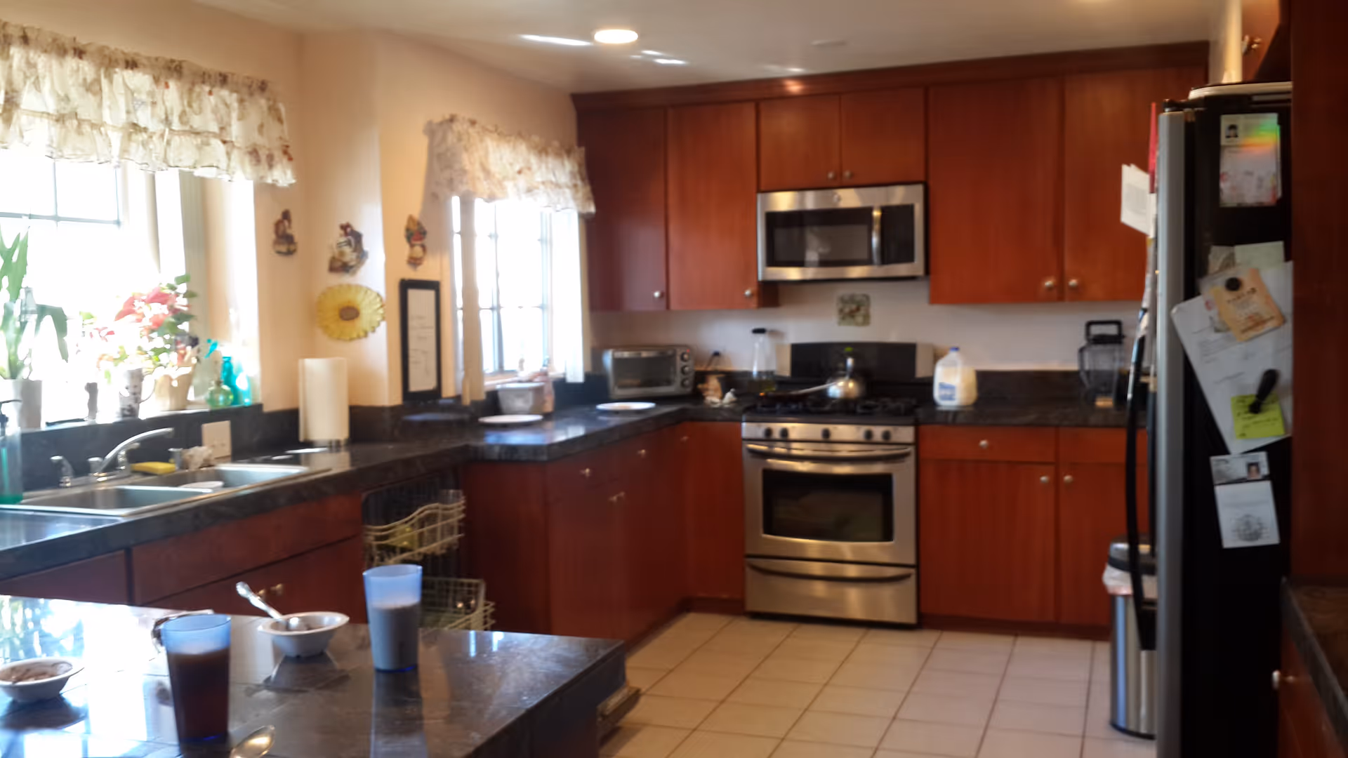 A kitchen with wooden cabinets, a stainless steel oven and microwave, a black countertop, and a refrigerator with papers and magnets on it. There are two windows with floral curtains letting in natural light. On the countertop, there are various items including a paper towel roll, a blender, a toaster oven, a gallon of milk, and some bowls and cups.