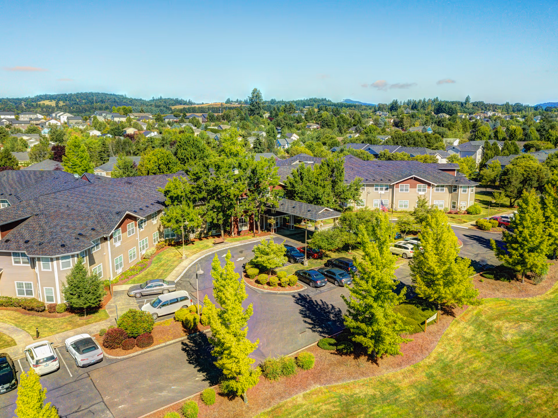 Aerial view of Vineyard Heights Assisted Living facility showing multiple two-story buildings with dark roofs surrounded by green trees and landscaped grounds. There is a parking lot with several cars and a covered entrance area. The background includes a suburban neighborhood and hills under a clear blue sky.