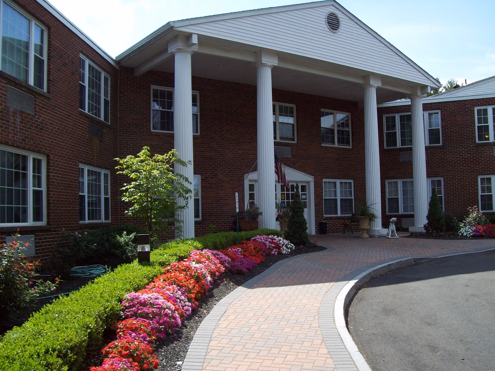 Brick senior living building with white columns, entryway, and colorful flower beds.