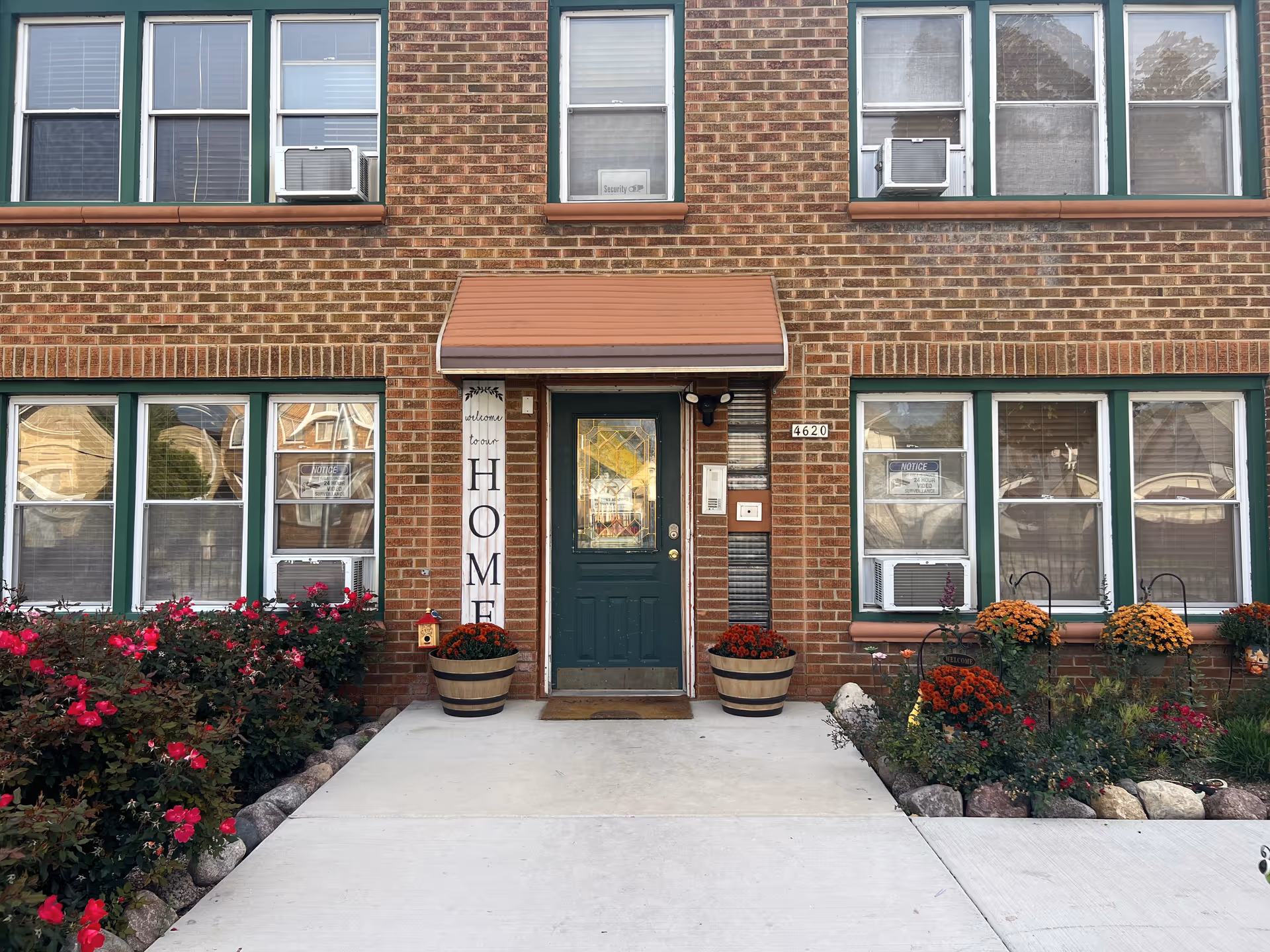 Brick building exterior with green-trimmed windows, entrance door, and flower-filled landscaping.