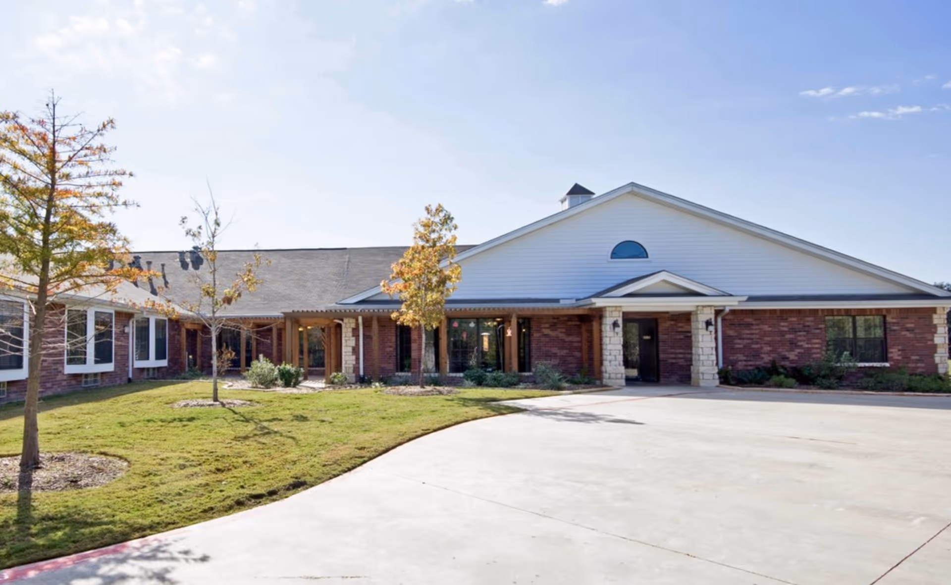 Front entrance of a single-story brick senior living facility with a curved driveway, lawn, and small trees.