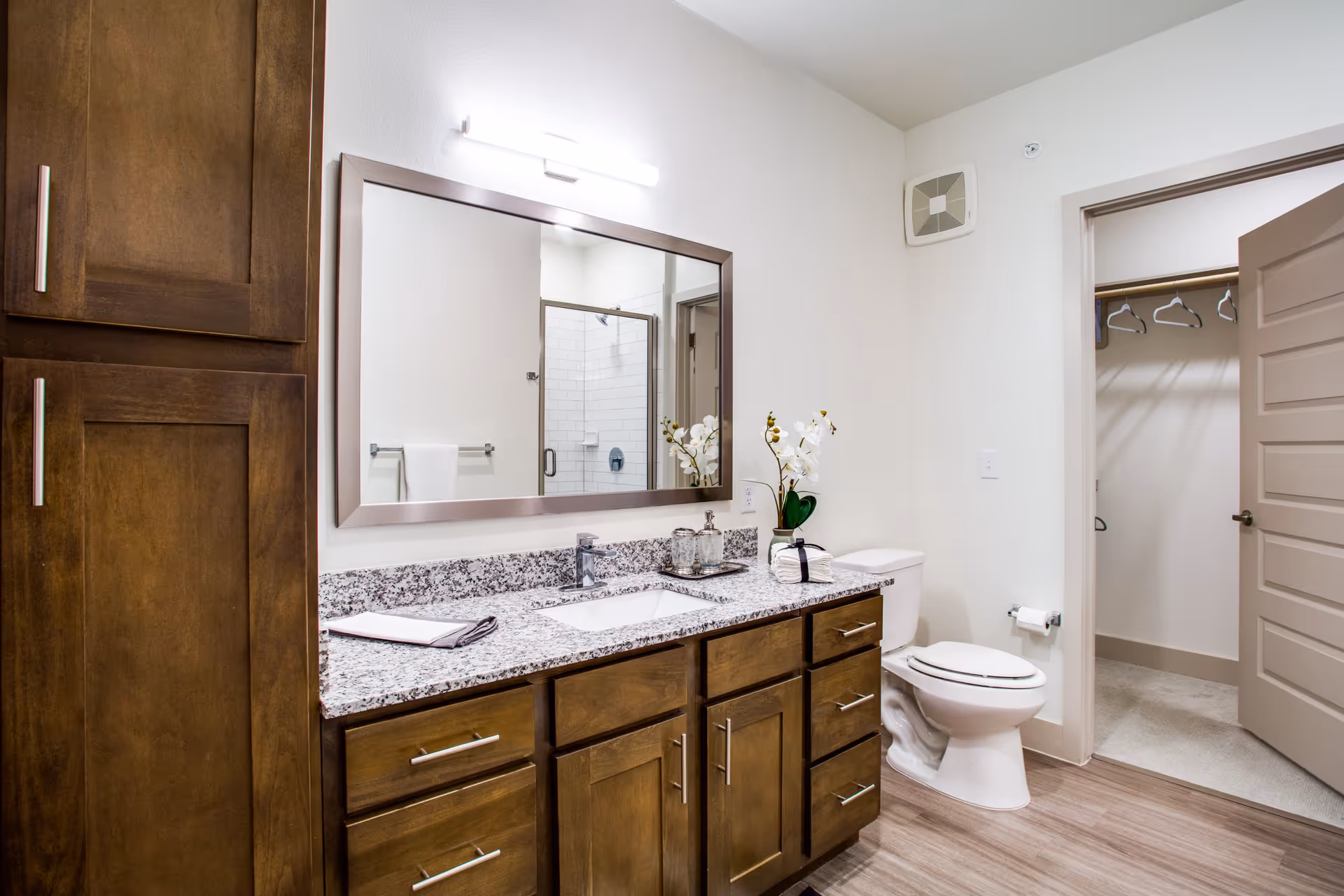 A modern bathroom with wooden cabinets and a granite countertop featuring a sink, a large rectangular mirror with a light above it, a white toilet, and an open door leading to a walk-in closet with hangers. The bathroom has light-colored walls and wood-style flooring.