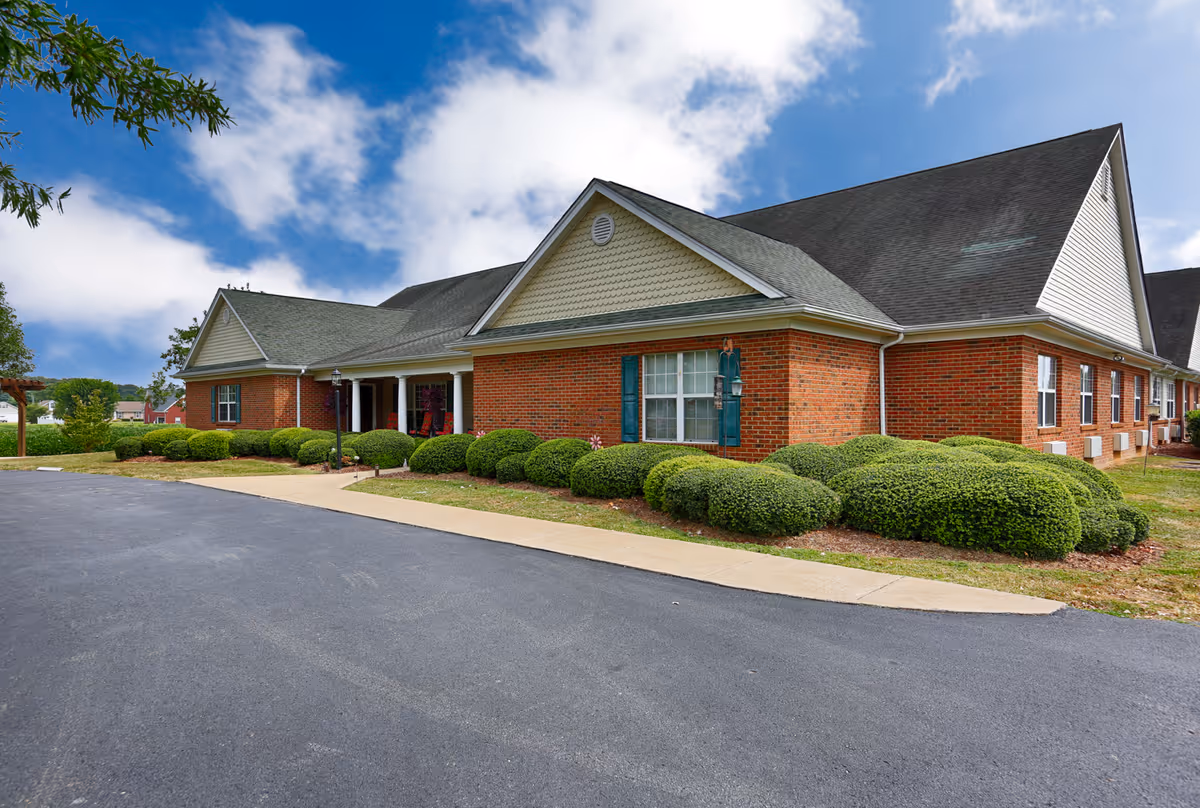 Exterior view of a single-story brick building with a gabled roof, surrounded by neatly trimmed bushes and a paved driveway under a partly cloudy sky.
