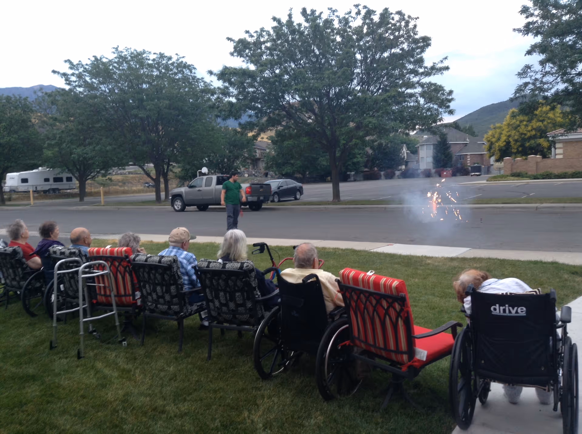 A group of elderly people, some in wheelchairs and others seated in chairs, are watching small fireworks being set off on a street in front of them. A man in a green shirt stands near the fireworks. The scene is outdoors with trees, parked cars, and houses in the background.