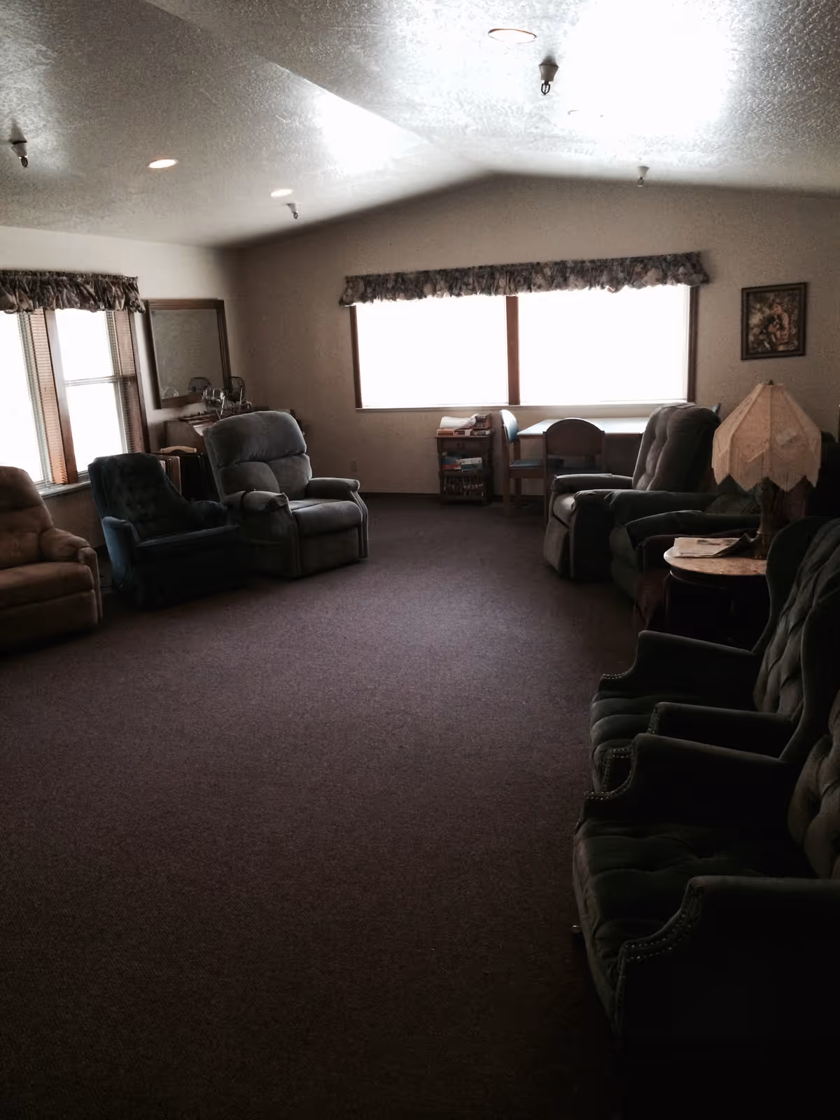 A dimly lit common room with several upholstered armchairs arranged along the walls. There are windows with floral valances letting in some light, a small table with chairs near the window, a side table with a lamp, and a framed picture on the wall.