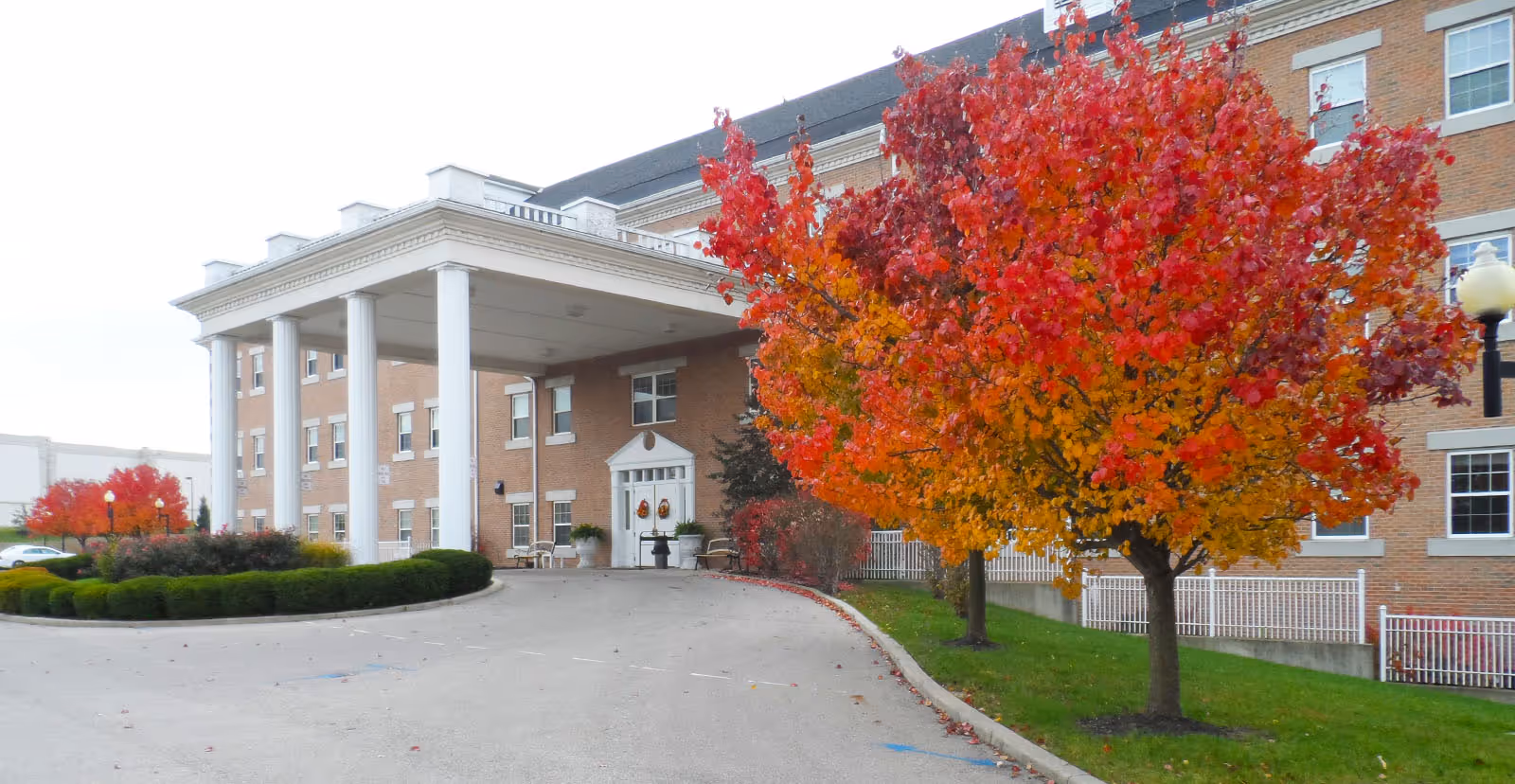 Exterior view of a multi-story brick building with white columns at the entrance. There are vibrant red and orange autumn trees lining the driveway leading to the entrance. The building has multiple windows and a white fence along the side.