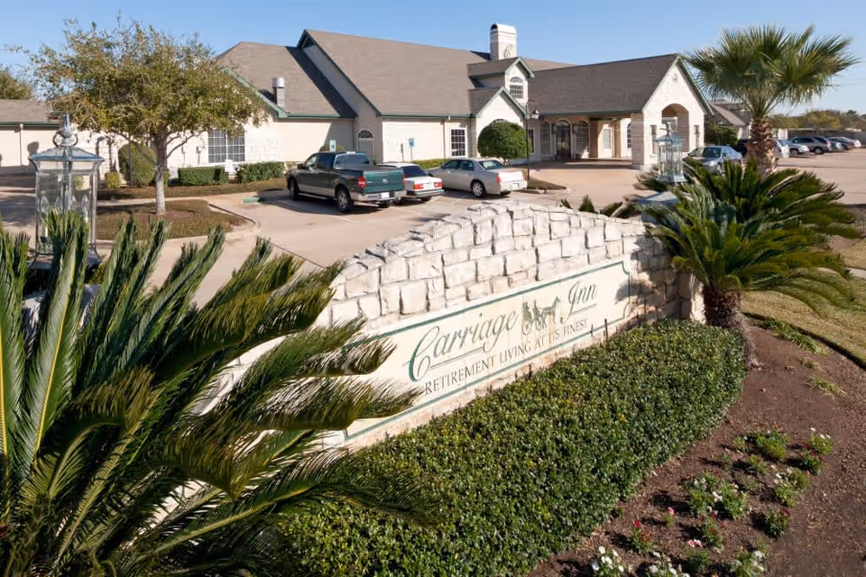Stone sign reading "Carriage Inn" with landscaped palms, flowers and a parking lot in front of the entrance to a single-story retirement facility.