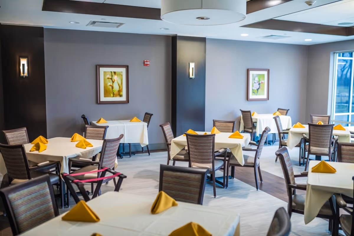 A dining room in a senior living facility with multiple tables covered in white tablecloths, each set with yellow folded napkins. The room has modern chairs, framed floral artwork on the walls, and large windows letting in natural light.