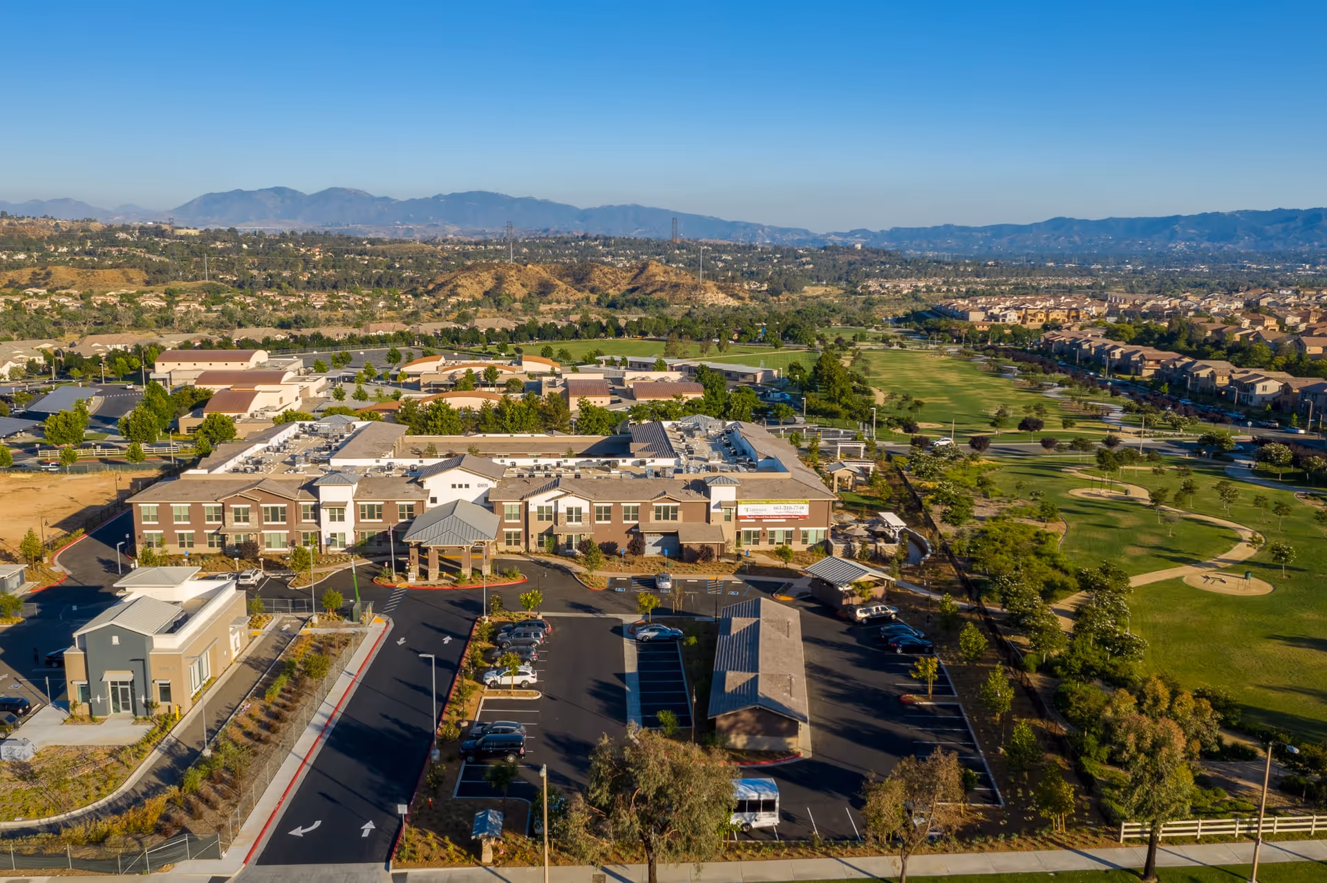Aerial view of Oakmont of Valencia senior living facility with a large building, parking lots, surrounding greenery, and a golf course adjacent to the property. Mountains are visible in the background under a clear blue sky.