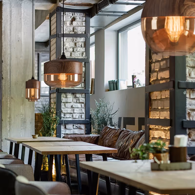 Modern indoor dining area with wooden tables, pendant lights, brick accents, and plants.