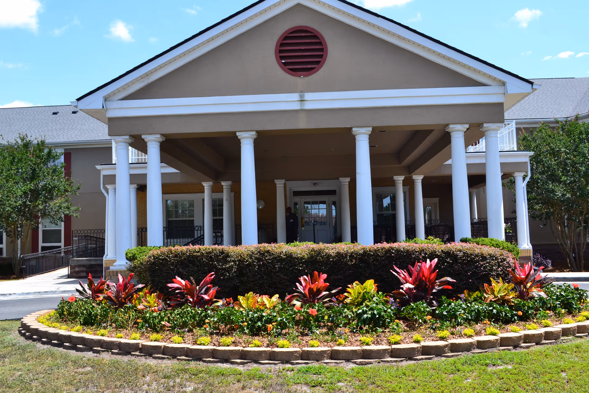 Covered front entrance of a residential building with white columns and a circular landscaped flower bed in front.
