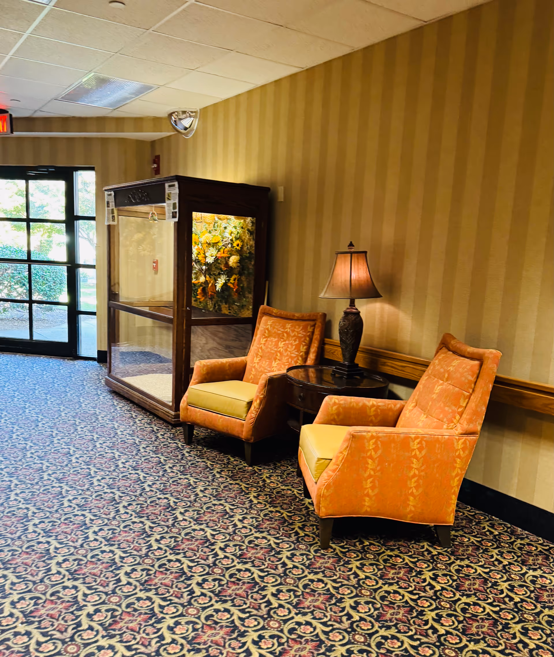 Orange upholstered chairs and a lamp sit beside a glass cabinet in a patterned lobby.