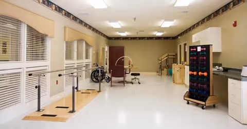 A spacious physical therapy room with parallel bars for walking exercises, a wheelchair, exercise equipment including colorful dumbbells on a rack, and a therapy chair. The room has large windows with blinds, light-colored walls, and a clean, bright floor.