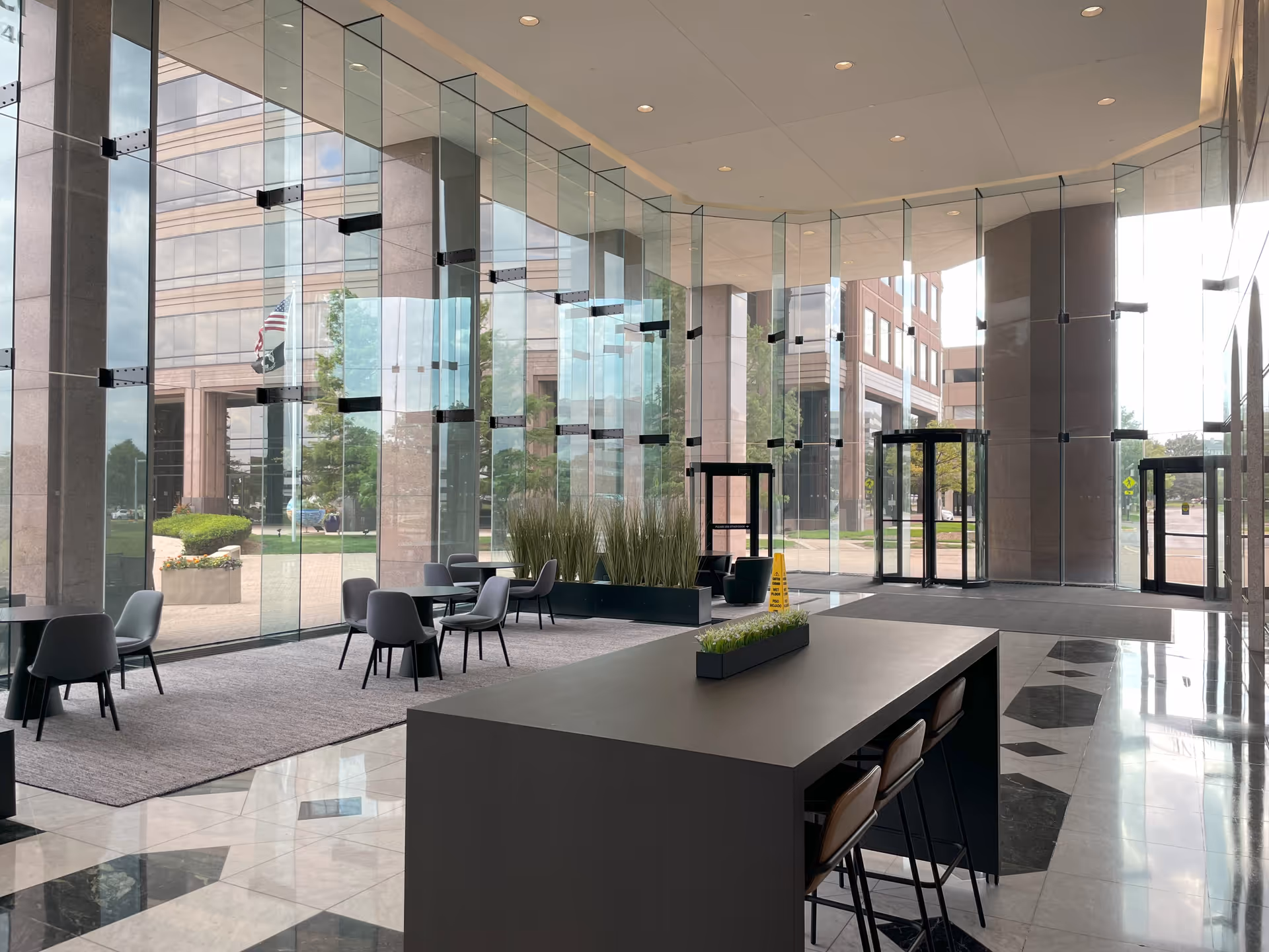 Modern glass-walled lobby with seating, a long reception desk, and planters.