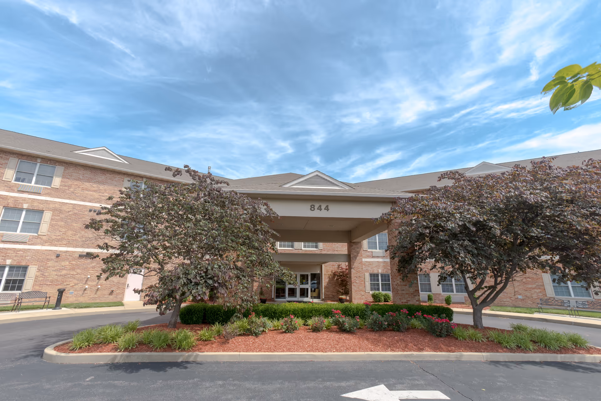 Front exterior view of a brick senior living facility building with the number 844 above the entrance. The entrance is covered by a portico supported by columns. There are two trees and landscaped flower beds with red mulch in front of the building. The sky is partly cloudy with blue and white clouds.