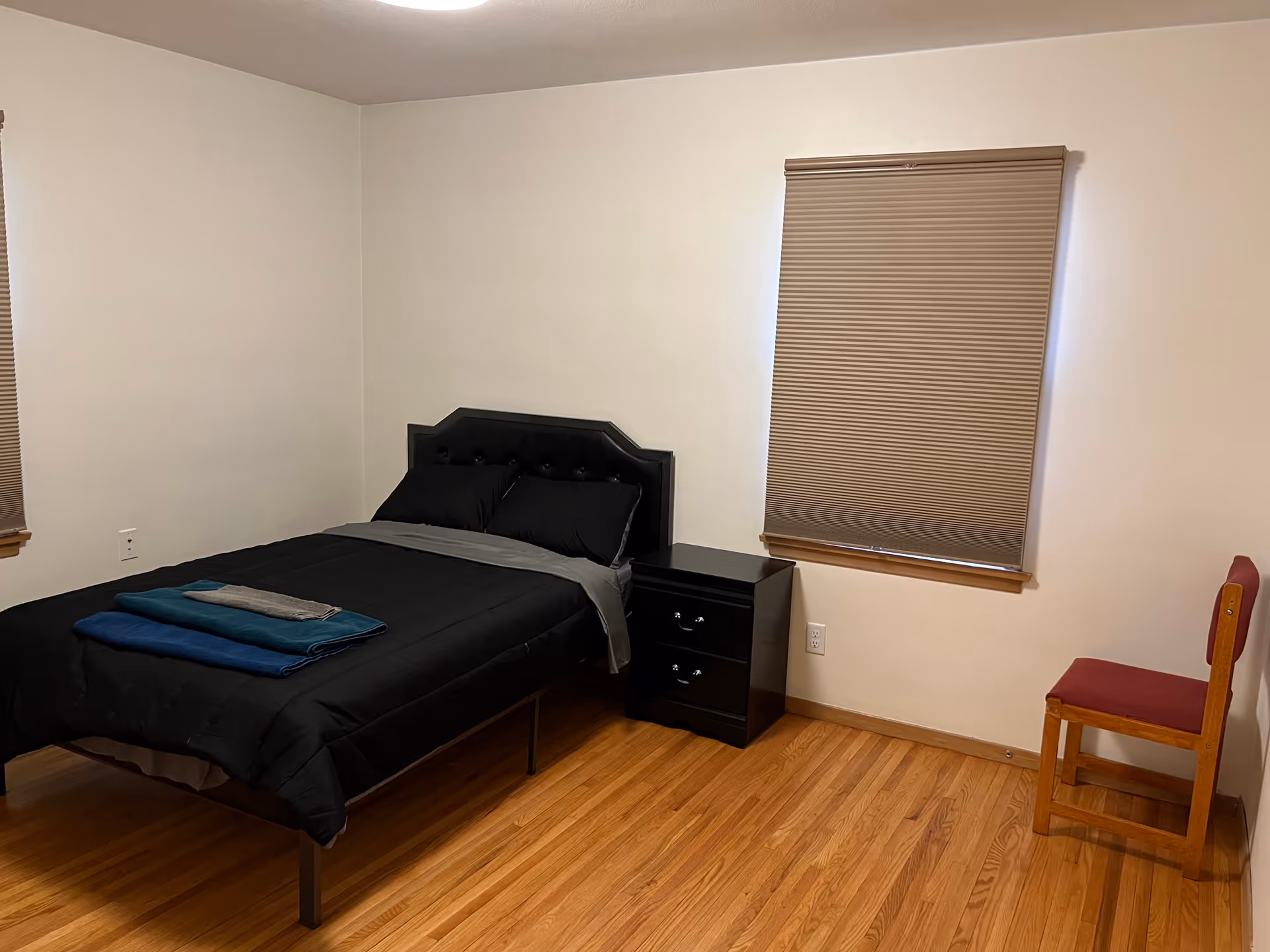 A furnished bedroom features a black bed, nightstand, chair, and blinds.