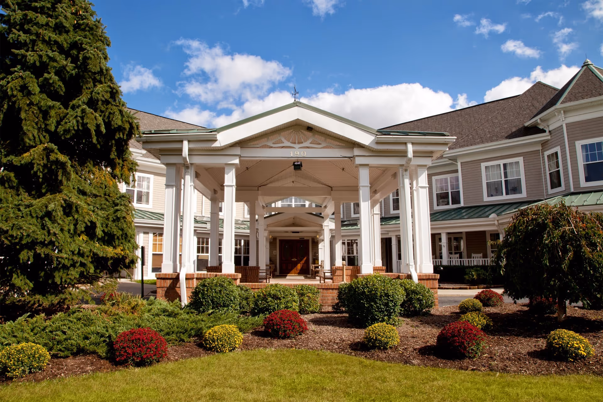 Front entrance of a senior living building with a covered porte-cochère, white columns, and landscaped shrubs and flowers.