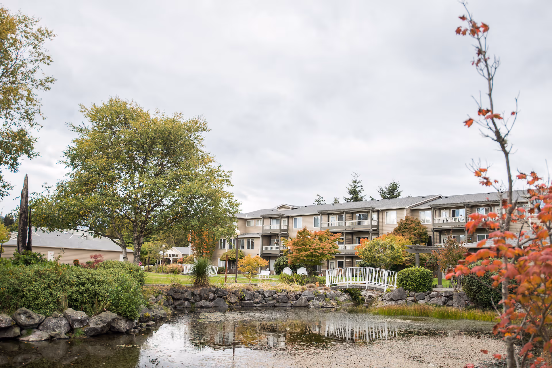 View of a senior living facility building named Village Concepts of Port Angeles - Park View Villas with a pond in the foreground, surrounded by rocks and greenery. There is a small white footbridge over the pond, trees with green and autumn-colored leaves, and a cloudy sky above.