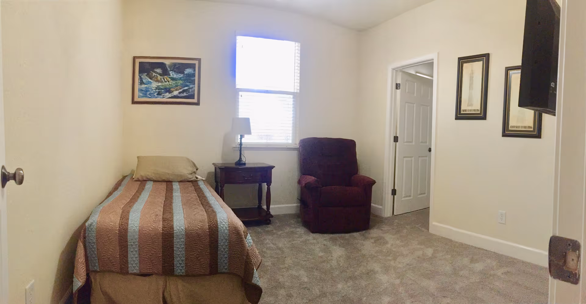 A small bedroom with a single bed covered with a striped brown and blue blanket, a beige pillow, a wooden nightstand with a lamp, a burgundy armchair, a window with blinds, and framed artwork on the walls.