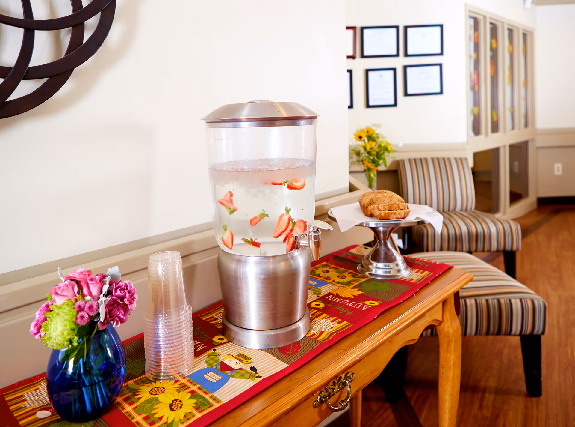 A refreshment table in a lounge area with a fruit-infused water dispenser, stacked plastic cups, a vase of flowers, pastries on a pedestal, and striped chairs in the background.