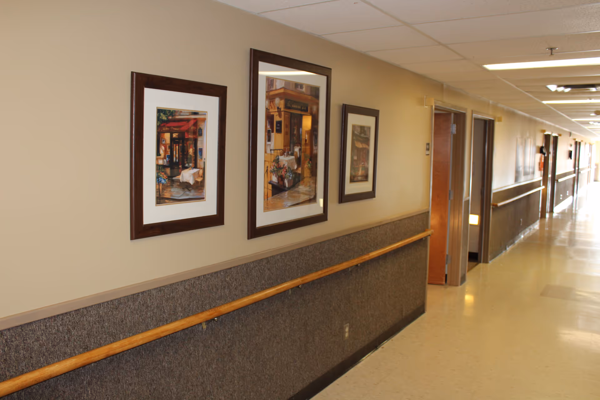 Long, well-lit corridor in a senior facility with framed pictures and a wall handrail.