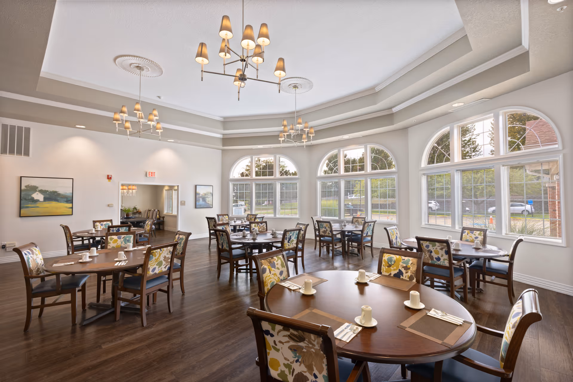 Bright dining room with round wooden tables, floral-upholstered chairs, chandeliers, and large arched windows.