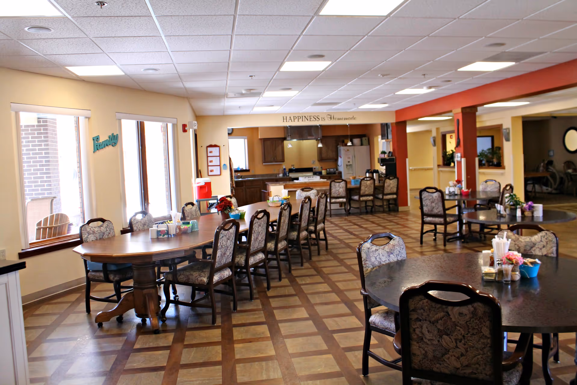 A spacious senior living dining area with multiple tables, chairs, and a serving kitchen in back.