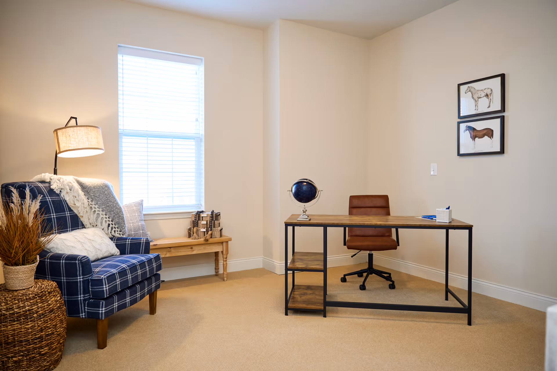 A cozy room with a blue plaid armchair draped with a white throw blanket and pillows, a wooden bench under a window with blinds, and a modern desk with a brown swivel chair. The desk has a small globe and a pen holder. Two framed pictures of horses hang on the beige wall.