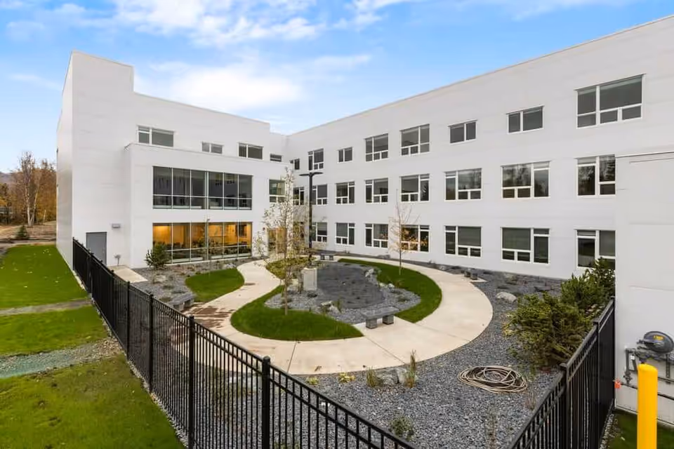 Outdoor courtyard area of a senior living facility with a circular walking path, small trees, and landscaped rocks, surrounded by a three-story white building with many windows and a black metal fence in the foreground.
