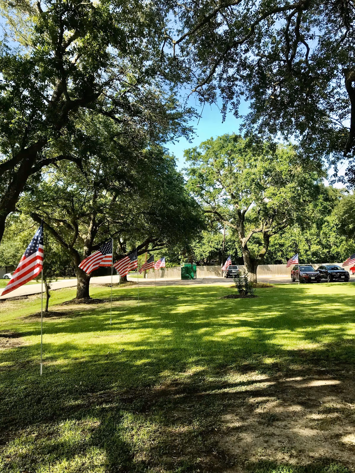 A grassy outdoor area with several American flags on poles lined up along a pathway. Large trees provide shade, and there are a few parked cars and a green dumpster visible in the background.